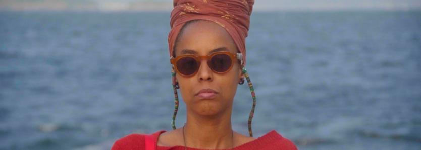 Participante do Seminário de Curadoria em Cinema e Audiovisual no Sesc Copacabana, Milena Mafredini, com cenário de praia ao fundo.