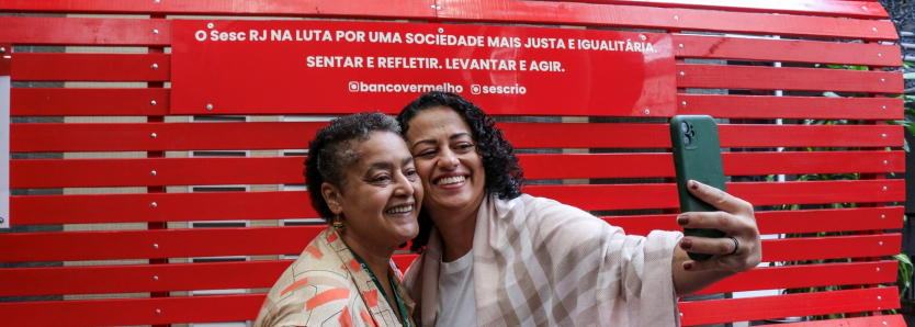 Duas mulheres sorriem enquanto tiram uma selfie em frente ao Banco Vermelho, campanha de conscientização contra a violência de gênero no Sesc RJ.